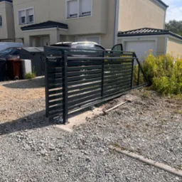 Nowoczesna, przesuwna brama wjazdowa z poziomych profili, zamontowana na posesji z domem w tle. Widoczny mechanizm jezdny i podłoże z kamiennego gruzu.