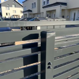 Nowoczesne, poziome ogrodzenie metalowe w kolorze grafitowym na tle domów jednorodzinnych. Widoczne elementy mocujące i żwirowe podłoże.