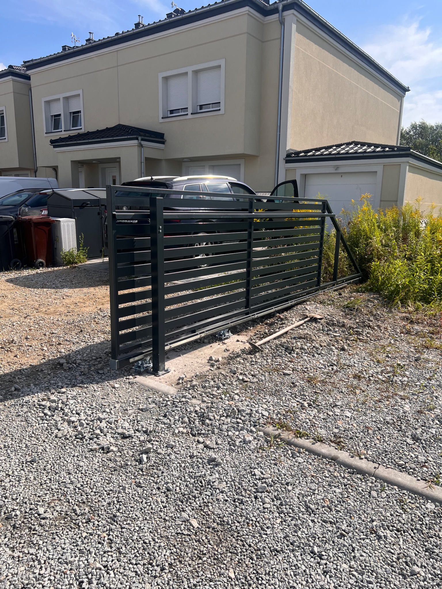 Nowoczesna, przesuwna brama wjazdowa z poziomych profili, zamontowana na posesji z domem w tle. Widoczny mechanizm jezdny i podłoże z kamiennego gruzu.