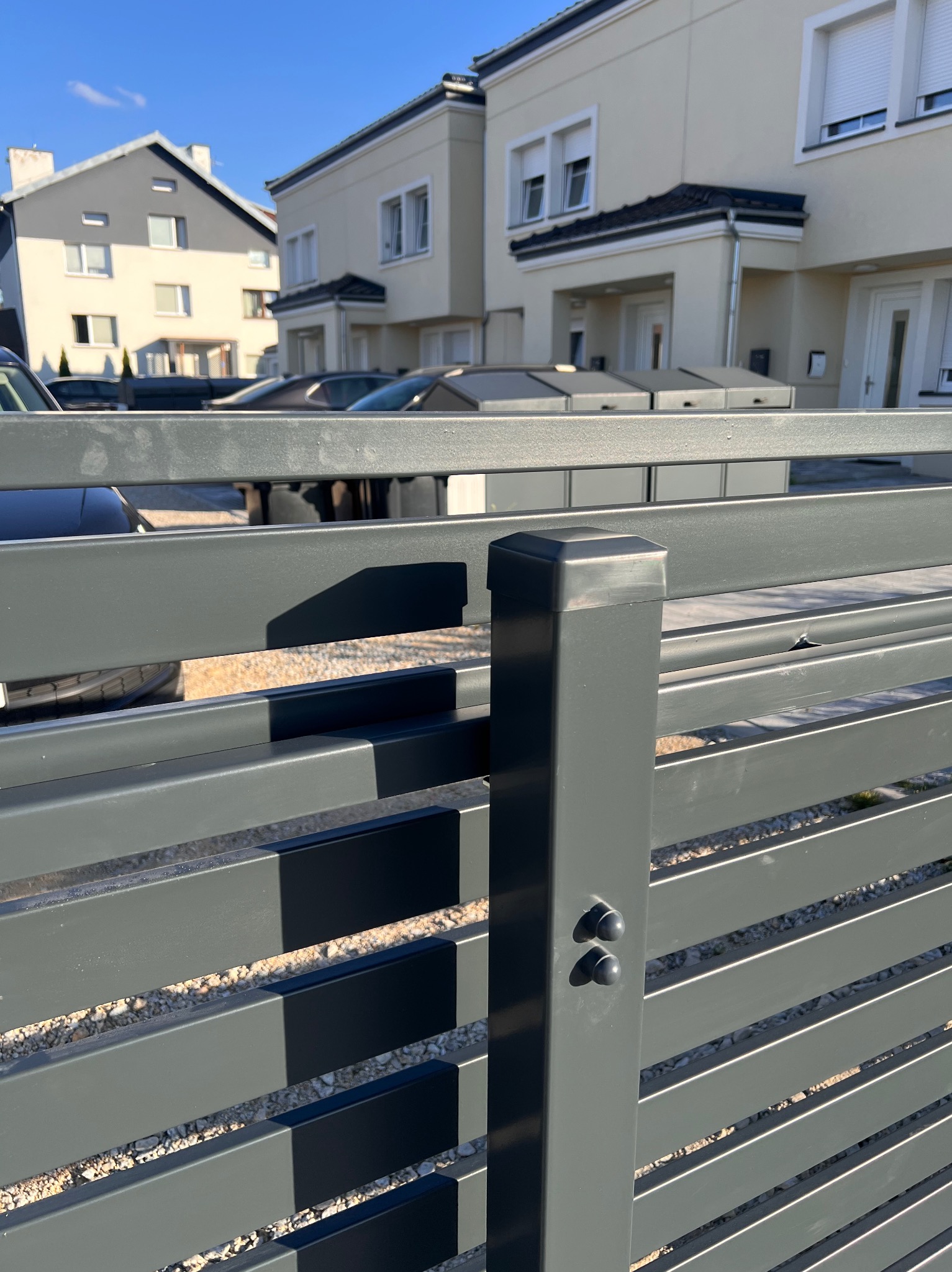 Nowoczesne, poziome ogrodzenie metalowe w kolorze grafitowym na tle domów jednorodzinnych. Widoczne elementy mocujące i żwirowe podłoże.