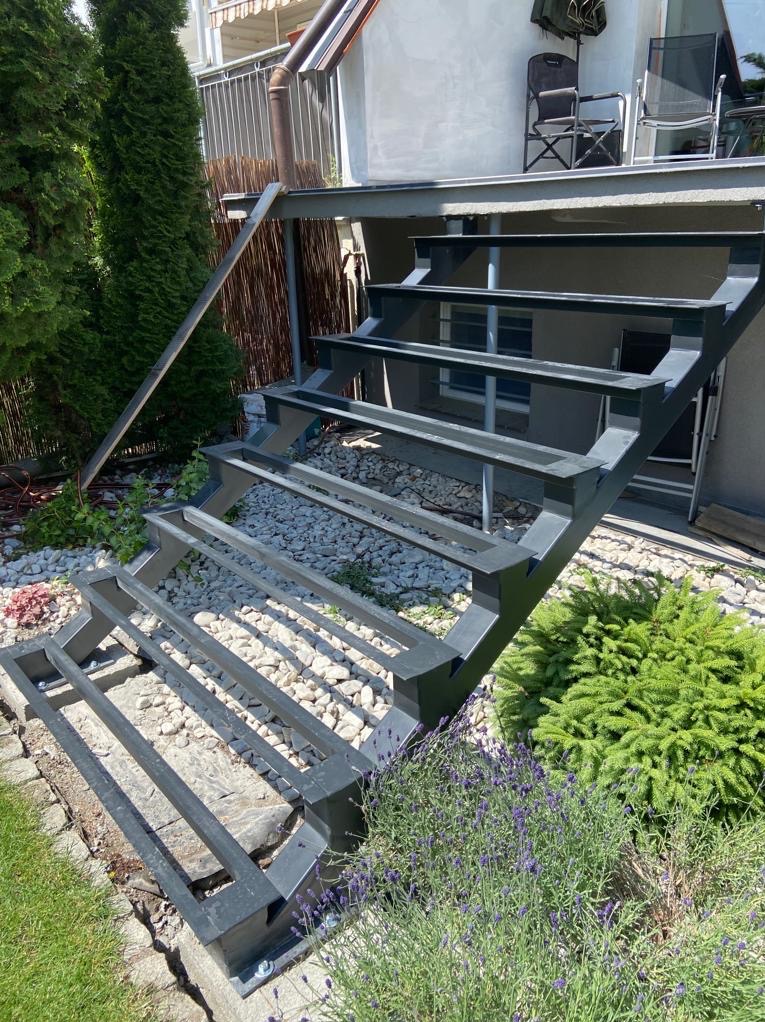 Metalowe schody zewnętrzne bez wypełnienia stopni, prowadzące na taras. Widoczne kamienie, trawa i roślinność. Szara konstrukcja schodów.