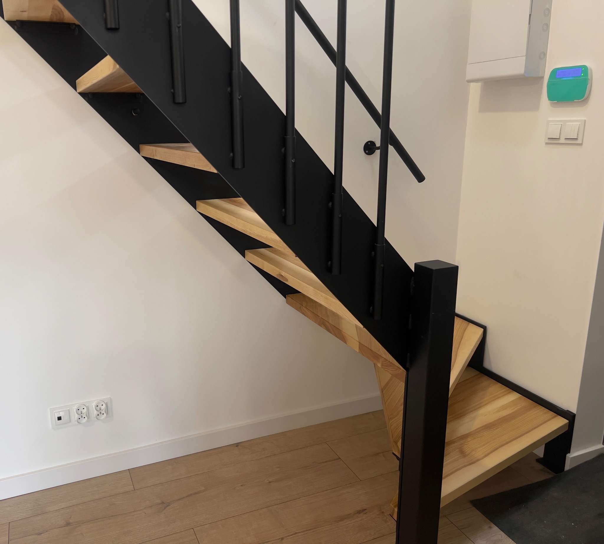 Nowoczesne schody z drewnianymi stopniami i czarną, metalową konstrukcją, widoczne z dołu, z elementami wykończenia wnętrza w tle.