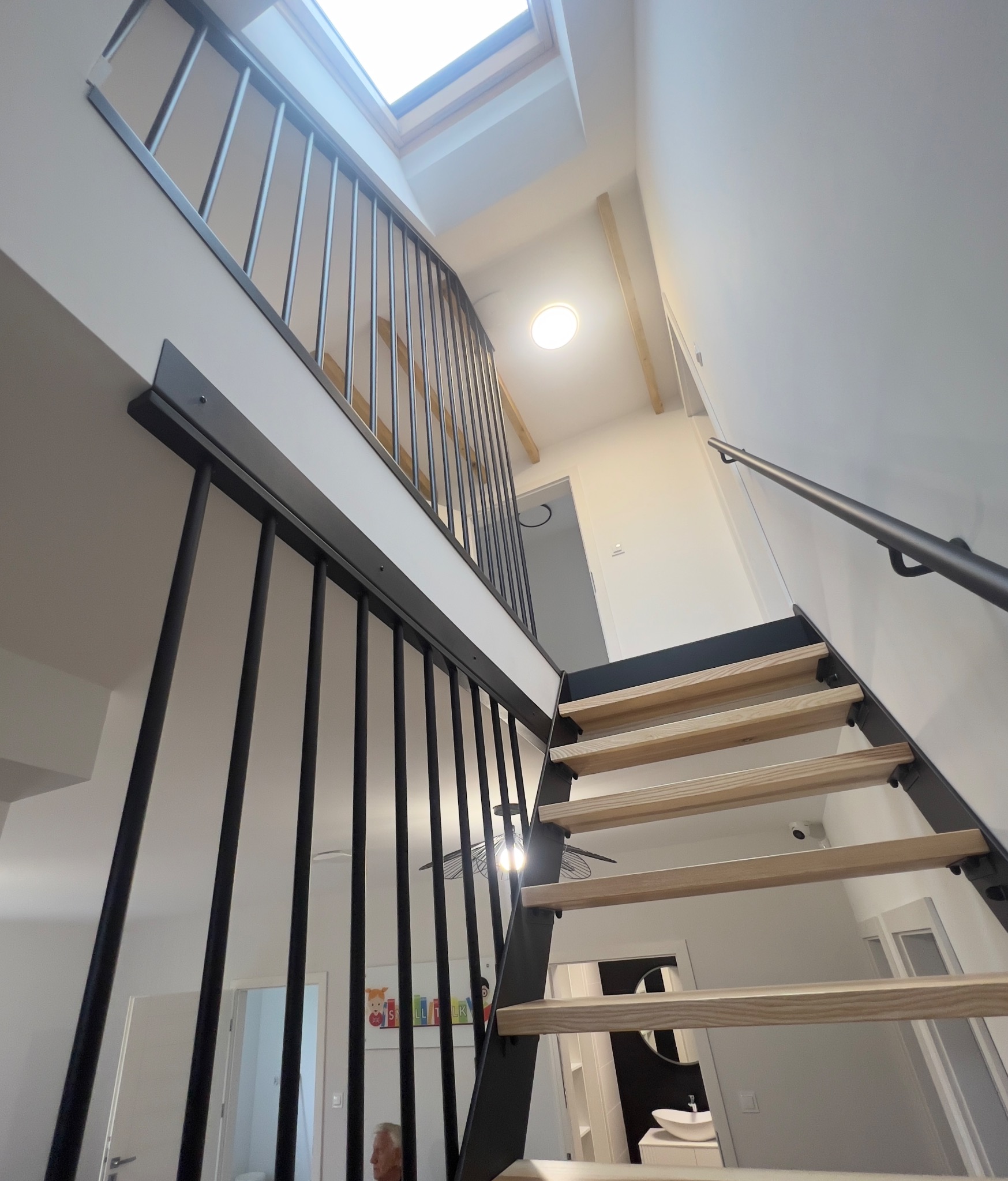 Nowoczesne schody z drewnianymi stopniami i czarną, metalową balustradą. Widok z dołu, w tle okno dachowe i oświetlenie sufitowe. Minimalistyczny design.