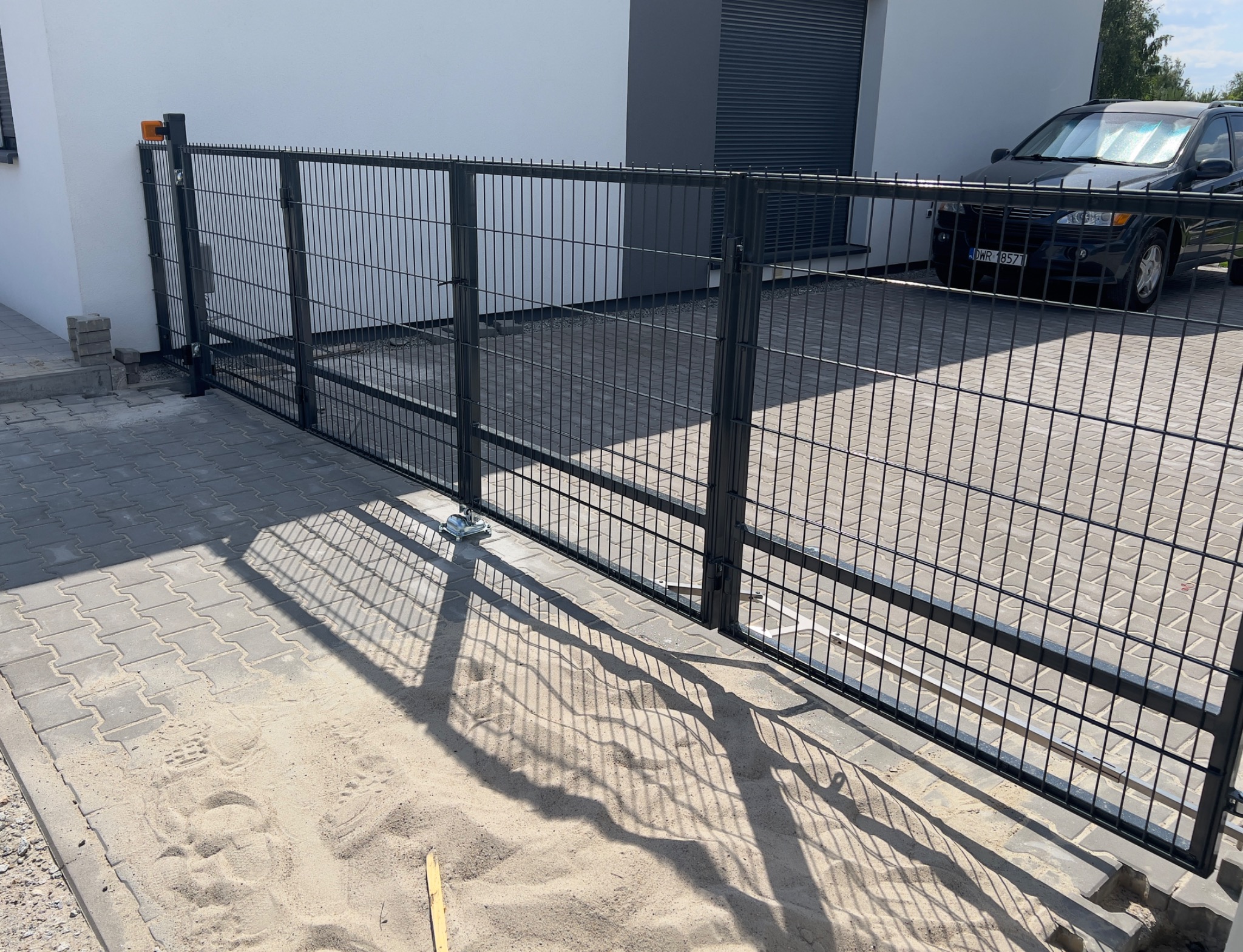 Nowoczesna, przesuwna brama z metalowej siatki w kolorze grafitowym, z widocznym automatem do bram. Na podjeździe ułożona kostka brukowa, w tle zaparkowany samochód.