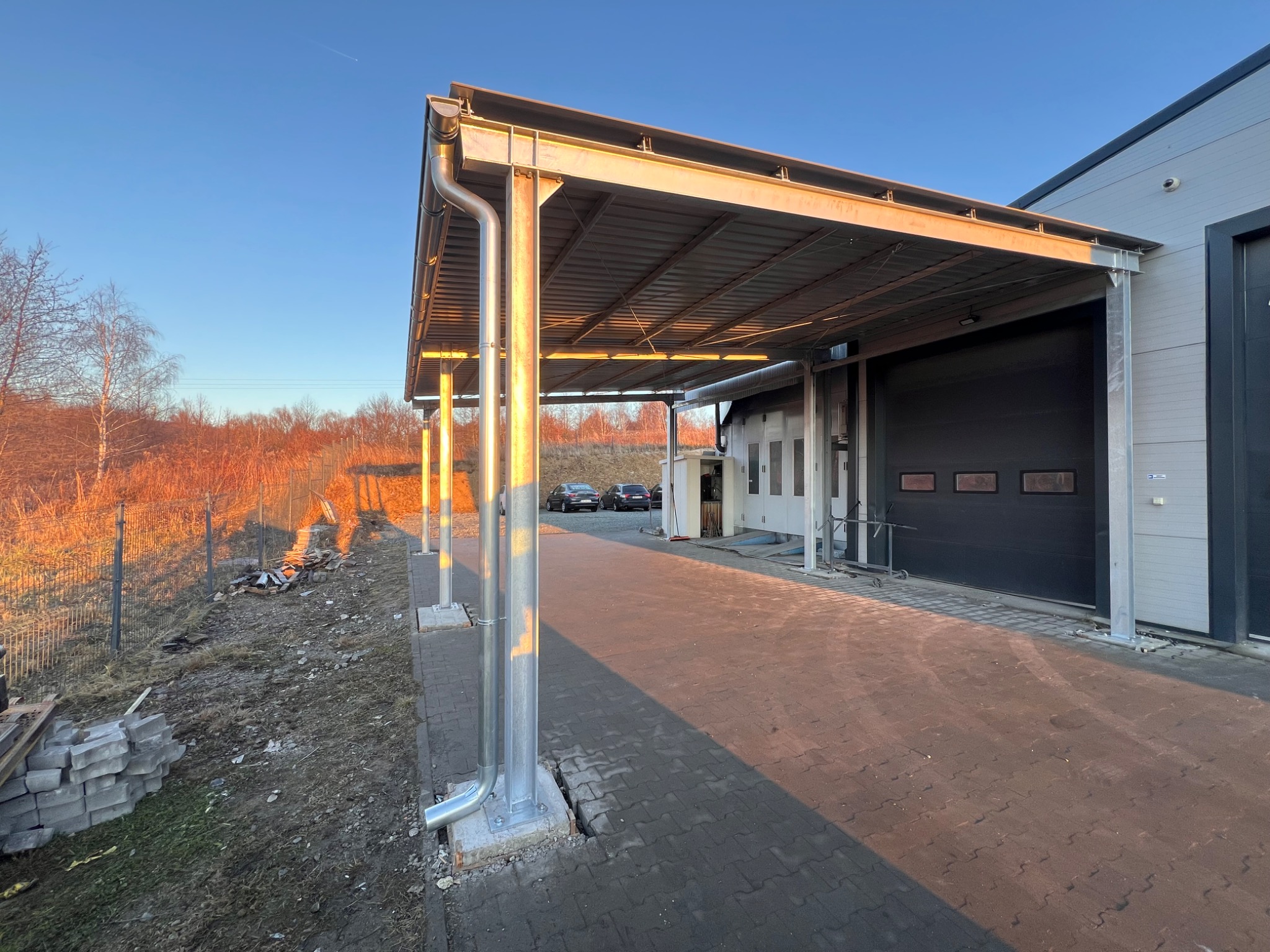 Metalowa konstrukcja zadaszenia przy budynku przemysłowym, widok z zewnątrz, w tle drzewa i błękitne niebo. Elementy konstrukcyjne w kolorze srebrnym.