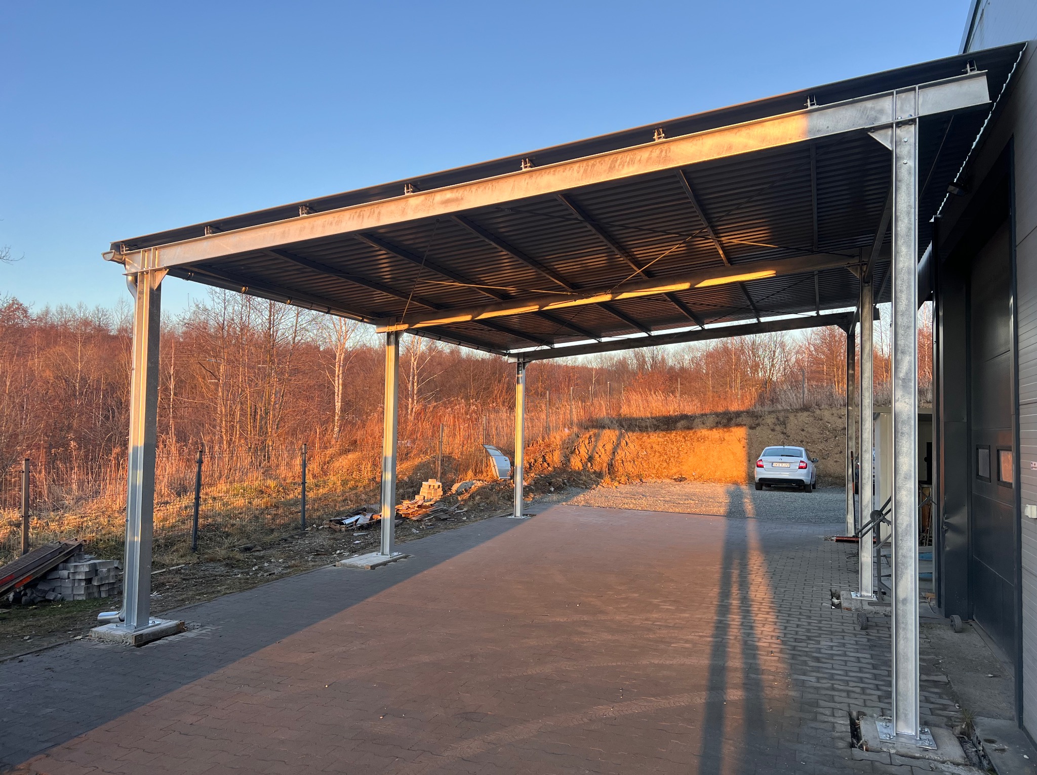 Metalowa konstrukcja z zadaszeniem, prawdopodobnie wiata, w promieniach zachodzącego słońca. W tle drzewa i zaparkowany samochód.