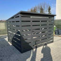 TechBlach - Nowoczesna, grafitowa konstrukcja z poziomych listew, prawdopodobnie na śmietnik, na tle błękitnego nieba i kostki brukowej. Widoczny cień osoby.