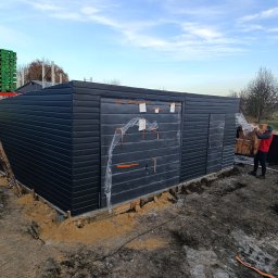 TechBlach - Montaż garażu blaszanego z poziomym panelem w kolorze grafitowym. Widoczne palety z materiałami budowlanymi i pracownik w trakcie pracy. Fundament w trakcie przygotowania.