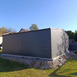 TechBlach - Szary garaż blaszany z poziomym wzorem paneli, widoczny na tle zielonego trawnika i błękitnego nieba. Ukończona konstrukcja na fundamencie, w tle budynki mieszkalne.