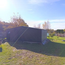 TechBlach - Nowoczesny, grafitowy garaż blaszany z bramą uchylną, usytuowany na trawniku w ogrodzie. Widoczny fragment osoby z taczką. Słoneczny dzień, błękitne niebo.