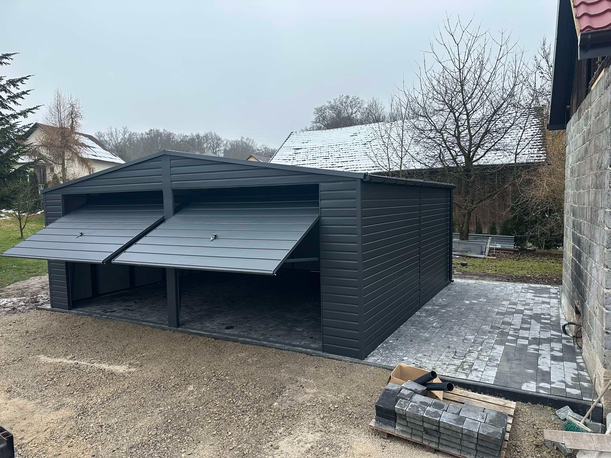 Dwustanowiskowy, grafitowy garaż blaszany z częściowo otwartymi bramami segmentowymi, na posesji z kostką brukową i widocznym śniegiem na dachu budynku w tle.