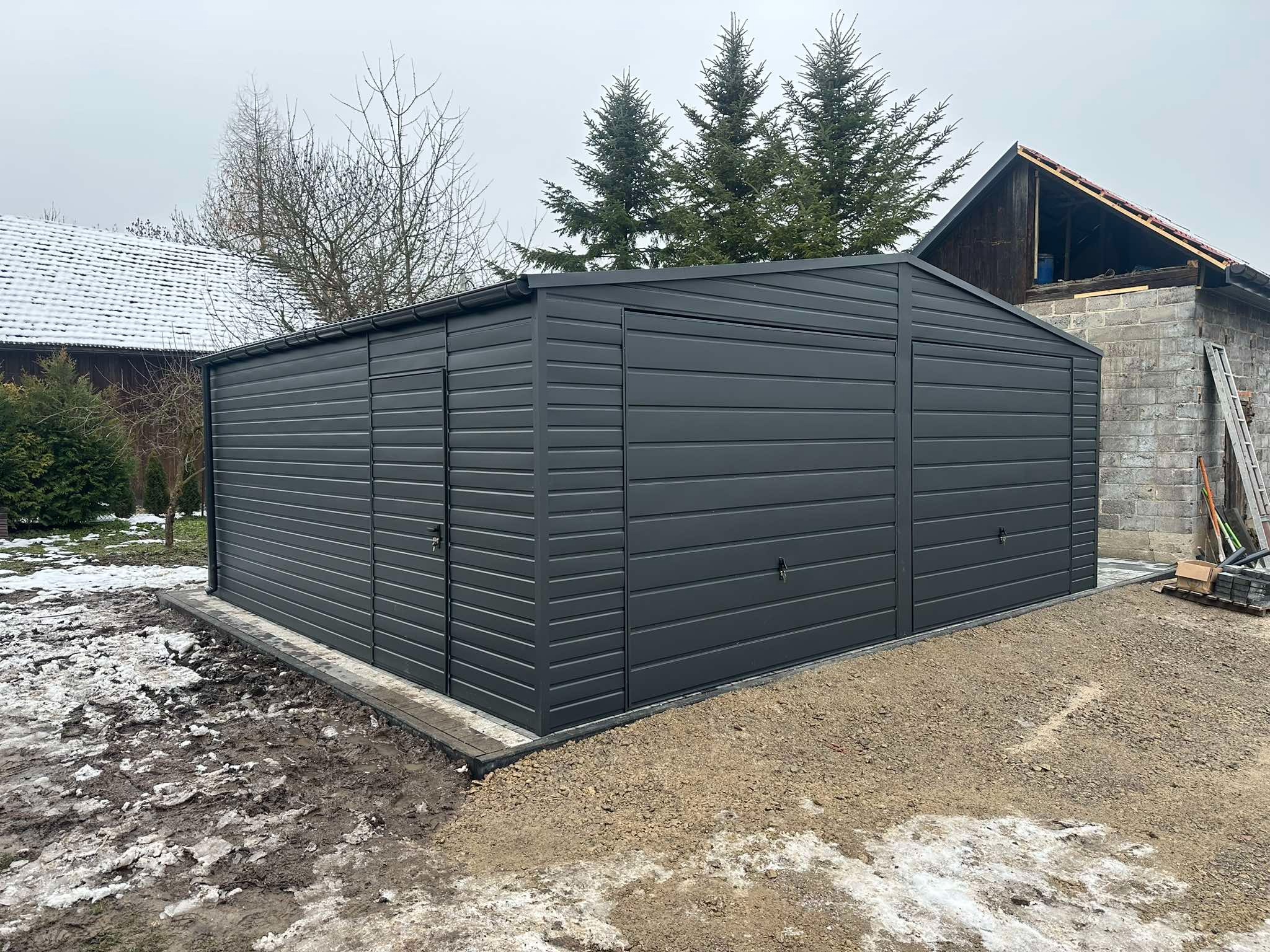 Nowoczesny, antracytowy garaż blaszany z trzema bramami, osadzony na betonowej podmurówce. W tle ośnieżony krajobraz i budynek w trakcie remontu dachu.