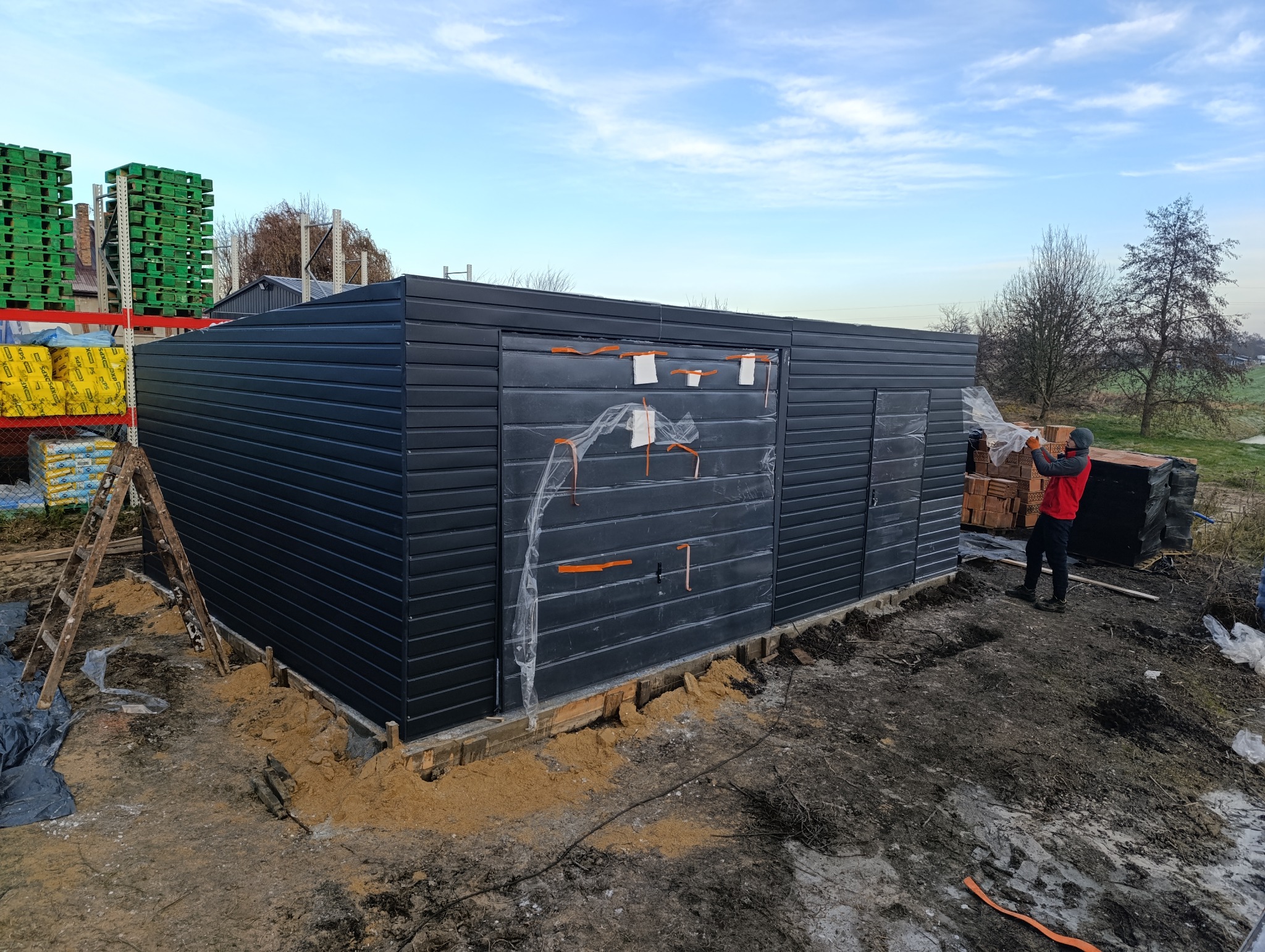 Montaż garażu blaszanego z poziomym panelem w kolorze grafitowym. Widoczne palety z materiałami budowlanymi i pracownik w trakcie pracy. Fundament w trakcie przygotowania.