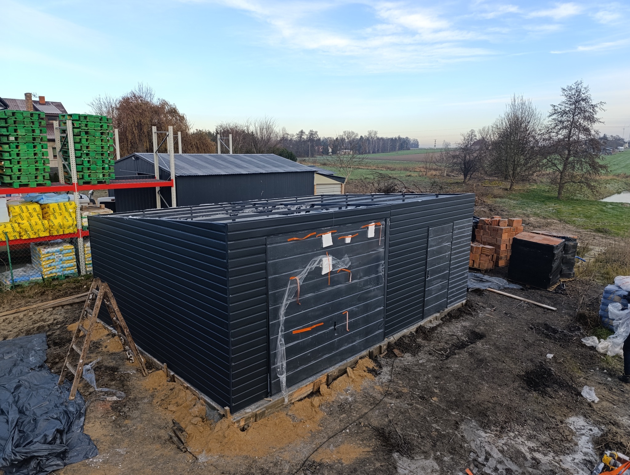 Montaż ciemnego garażu blaszanego z widocznymi elementami konstrukcyjnymi i przygotowanym podłożem, na tle krajobrazu wiejskiego.