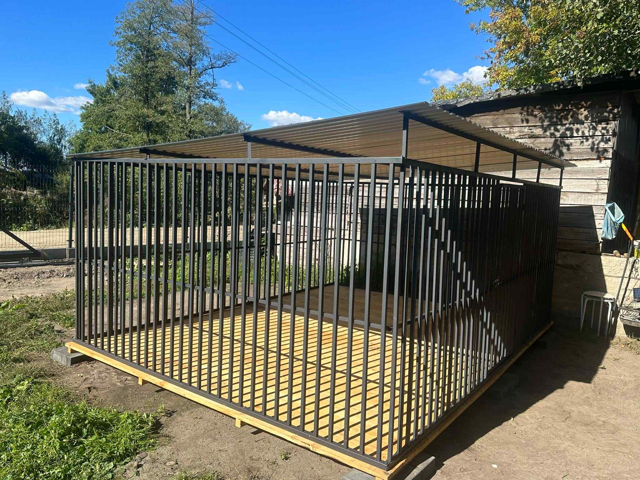 Metalowa konstrukcja z zadaszeniem, prawdopodobnie kojec dla psa, z drewnianą podłogą, na zewnątrz, w tle drzewa i budynek gospodarczy.