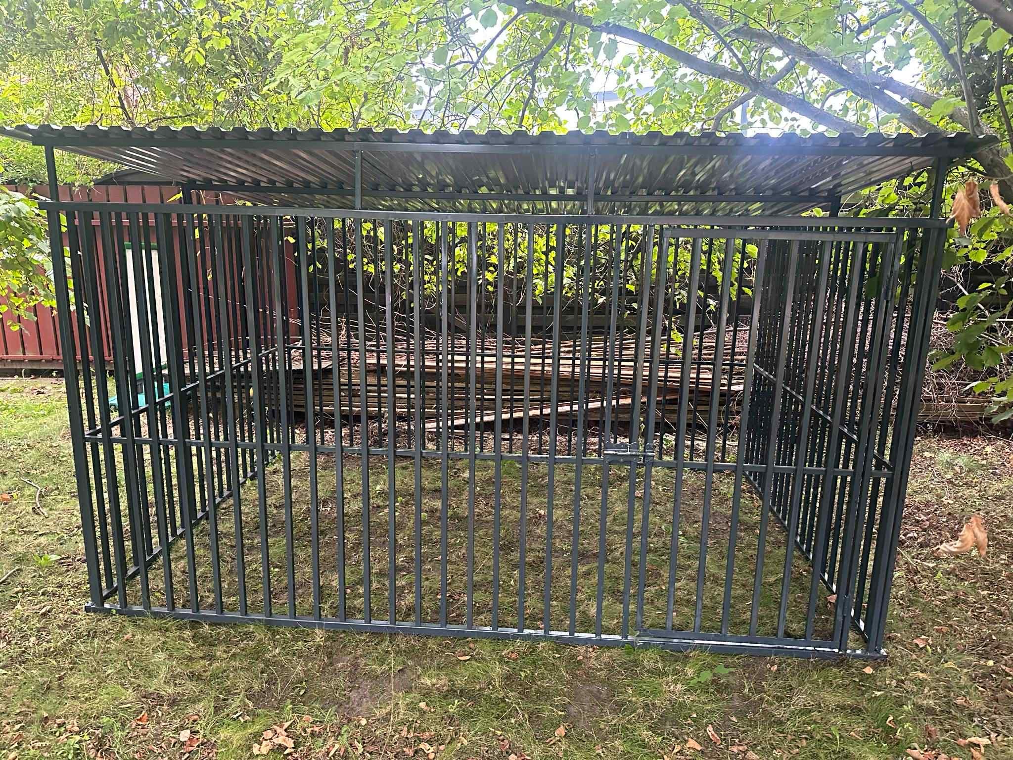 Metalowa konstrukcja z zadaszeniem z blachy falistej, przypominająca klatkę lub kojec, ustawiona na trawie pod drzewami. Widoczne elementy spawane i mocowania.