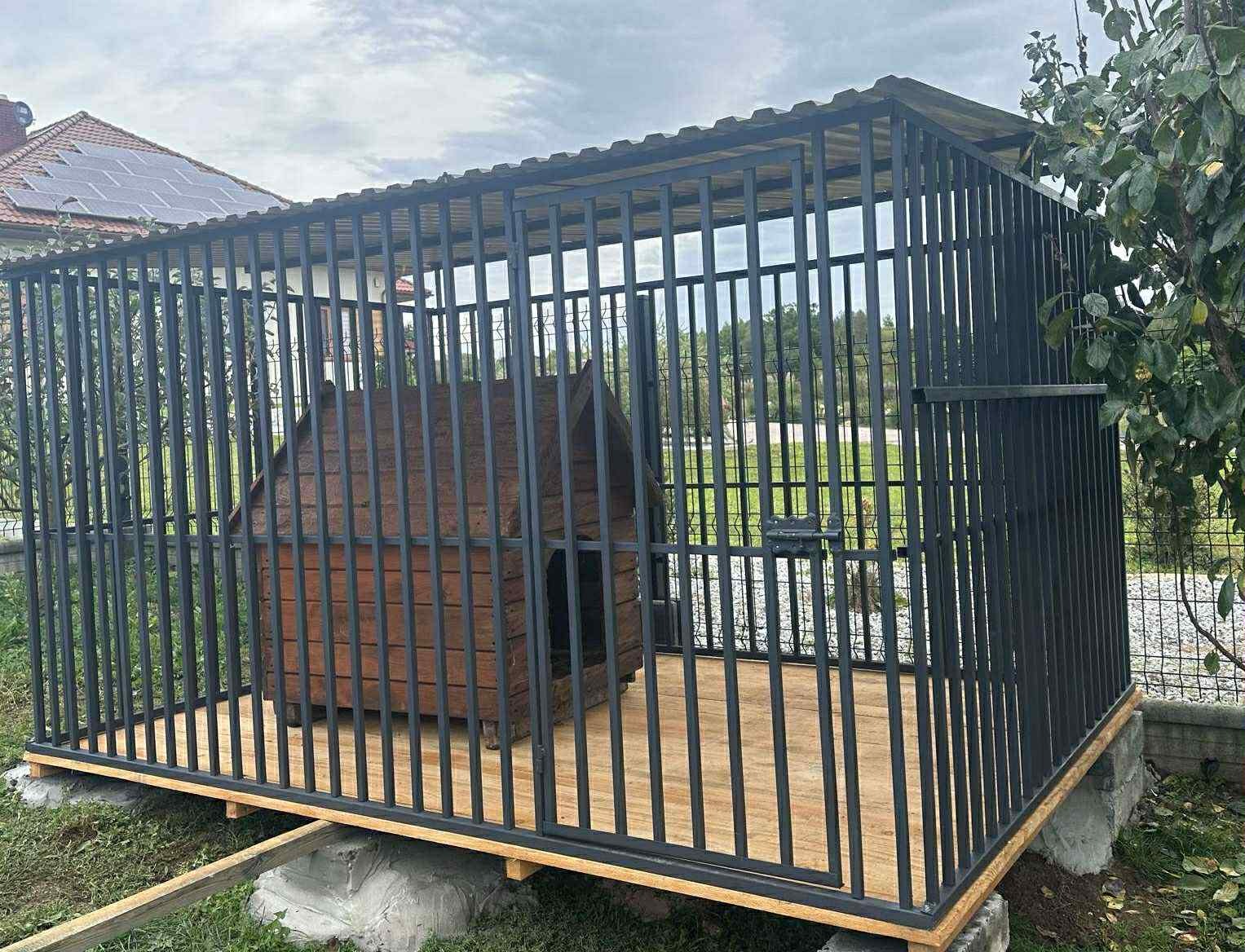 Metalowa konstrukcja zadaszona, z drewnianą budą w środku, na betonowej podstawie. Widoczne drzewa i dom z panelami słonecznymi w tle.