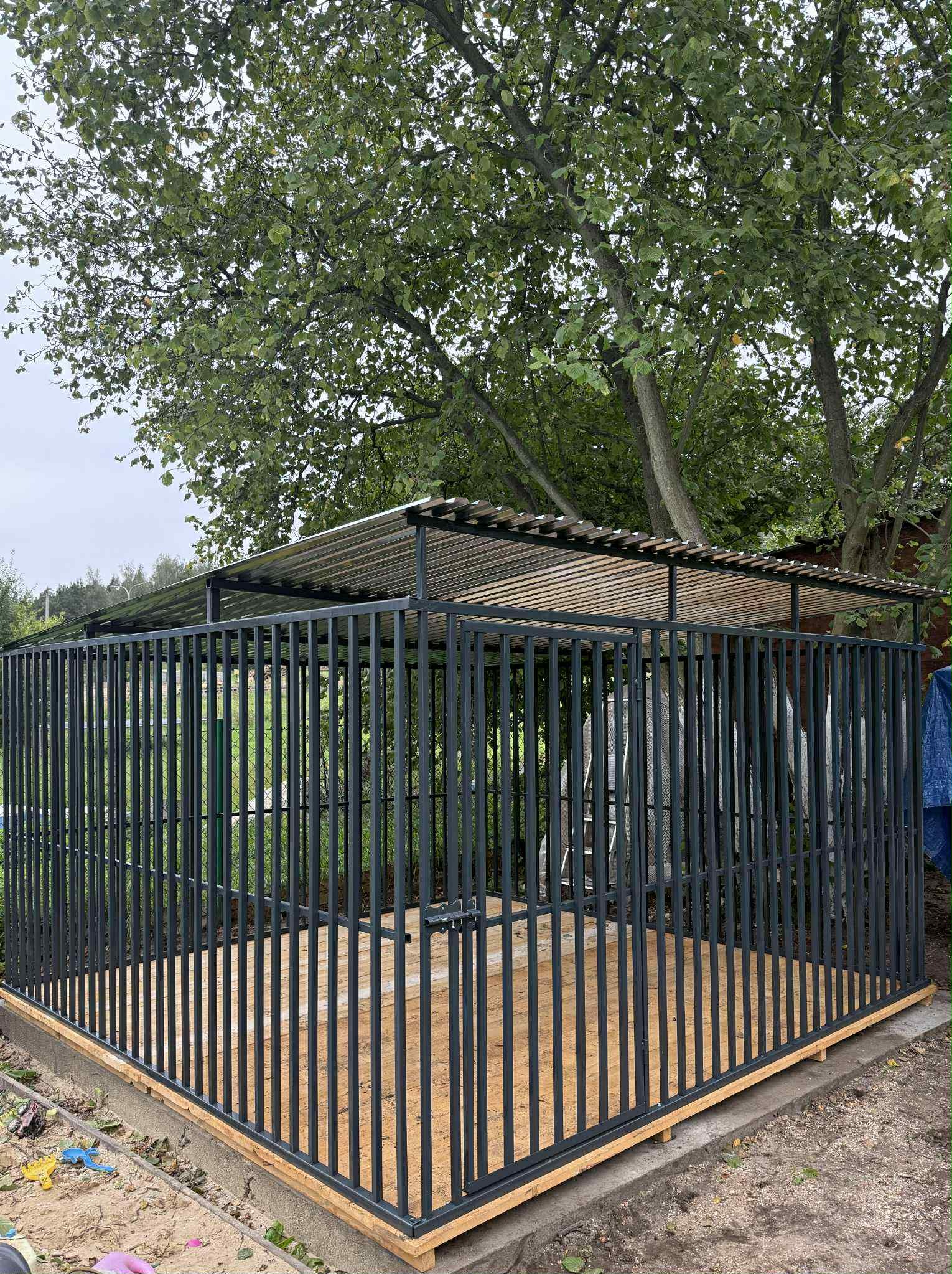 Metalowa konstrukcja z zadaszeniem na drewnianej podłodze, otoczona zielenią. Prostopadłościenne ujęcie prezentuje detale konstrukcyjne i materiały.