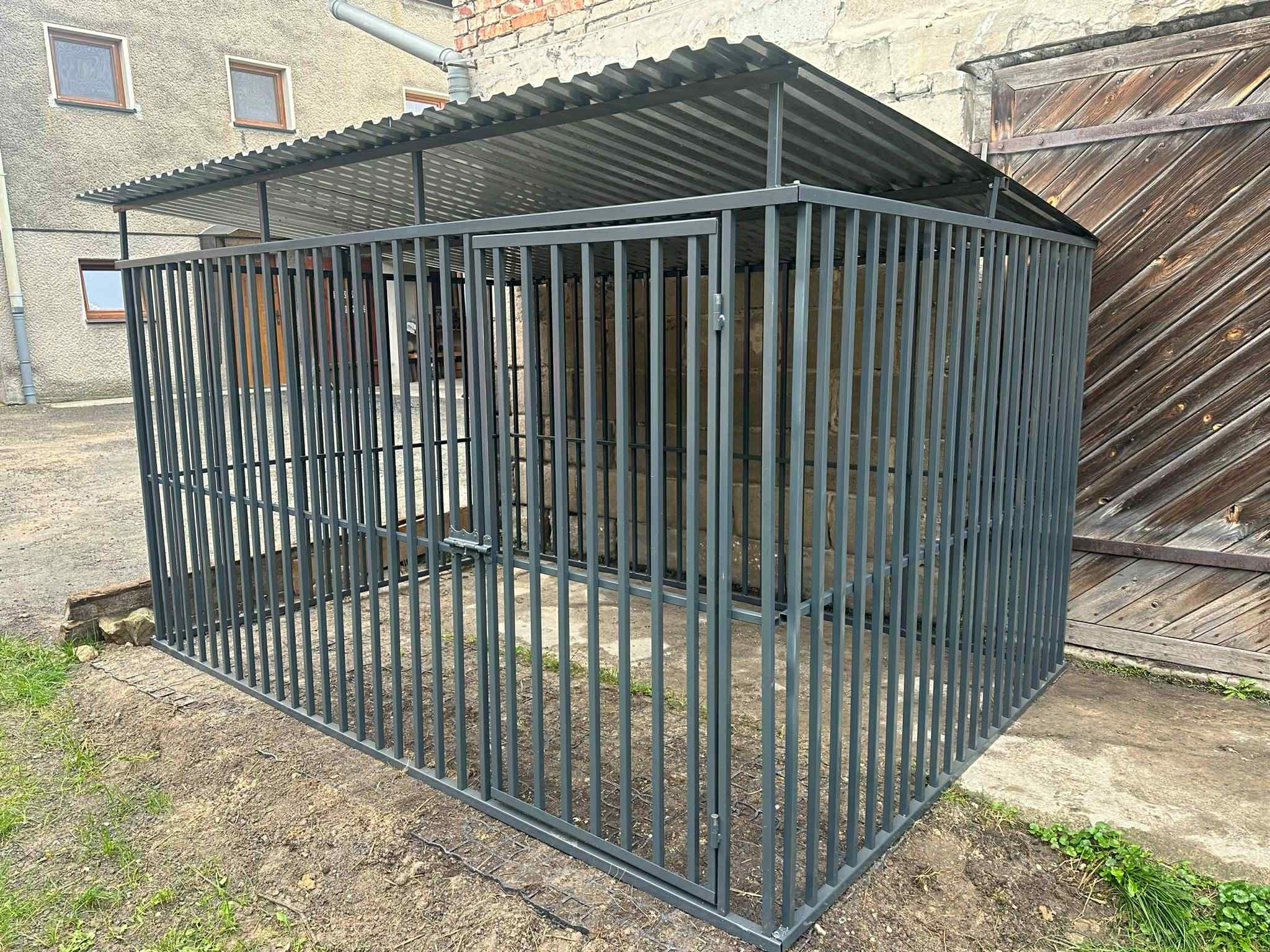 Metalowa konstrukcja z zadaszeniem w Dobczycach. Szara, pionowa kratownica z otwartymi drzwiami, zadaszona blachą falistą. Tło: budynek z cegły i drewniane wrota.