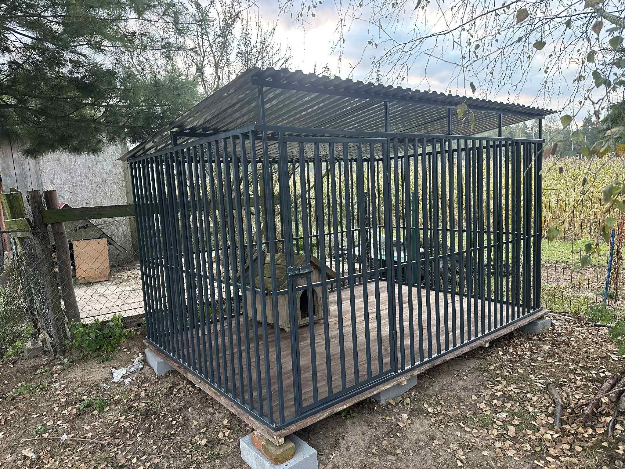 Metalowa konstrukcja z zadaszeniem i drewnianą podłogą, prawdopodobnie kojec dla psa, z budą w środku, umieszczona na betonowych bloczkach na zewnątrz.