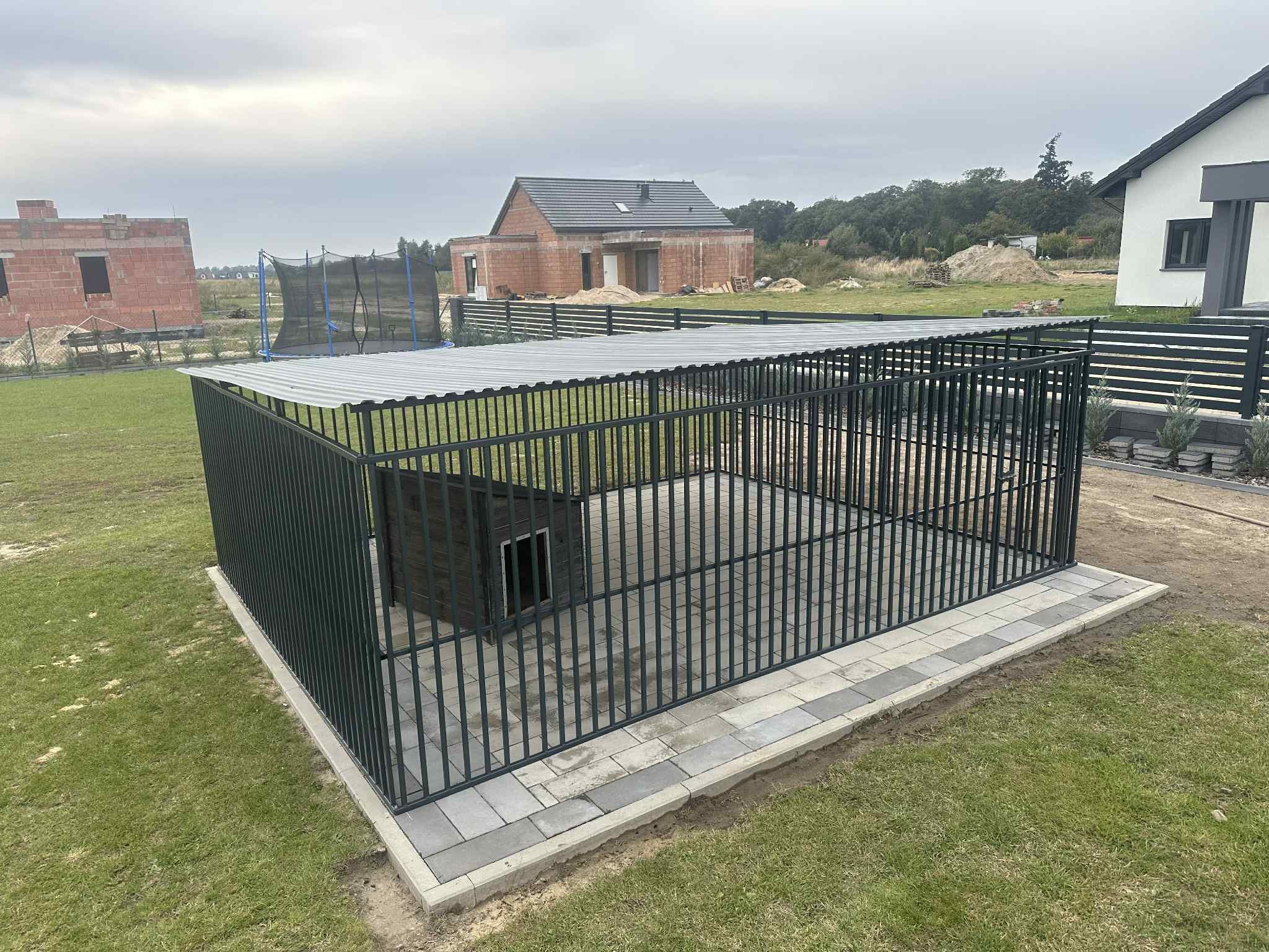 Metalowa konstrukcja z zadaszeniem i budą dla psa na posesji. Solidne wykonanie, widoczne detale spawów. Tło: dom w budowie, trawnik i ogrodzenie.