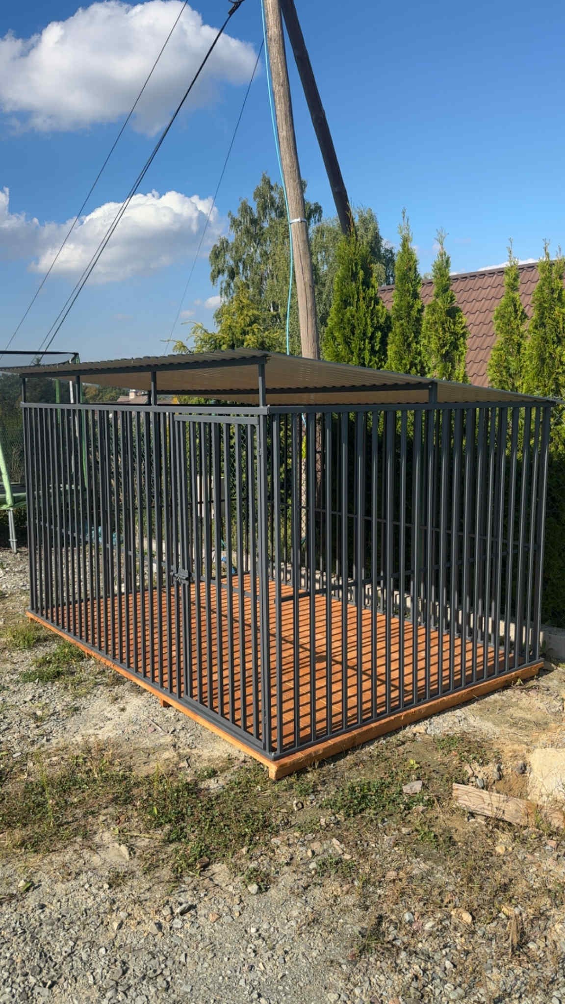 Metalowa konstrukcja z zadaszeniem i drewnianą podłogą na zewnątrz, otoczona zielenią i błękitnym niebem. Widoczne słupy energetyczne w tle.