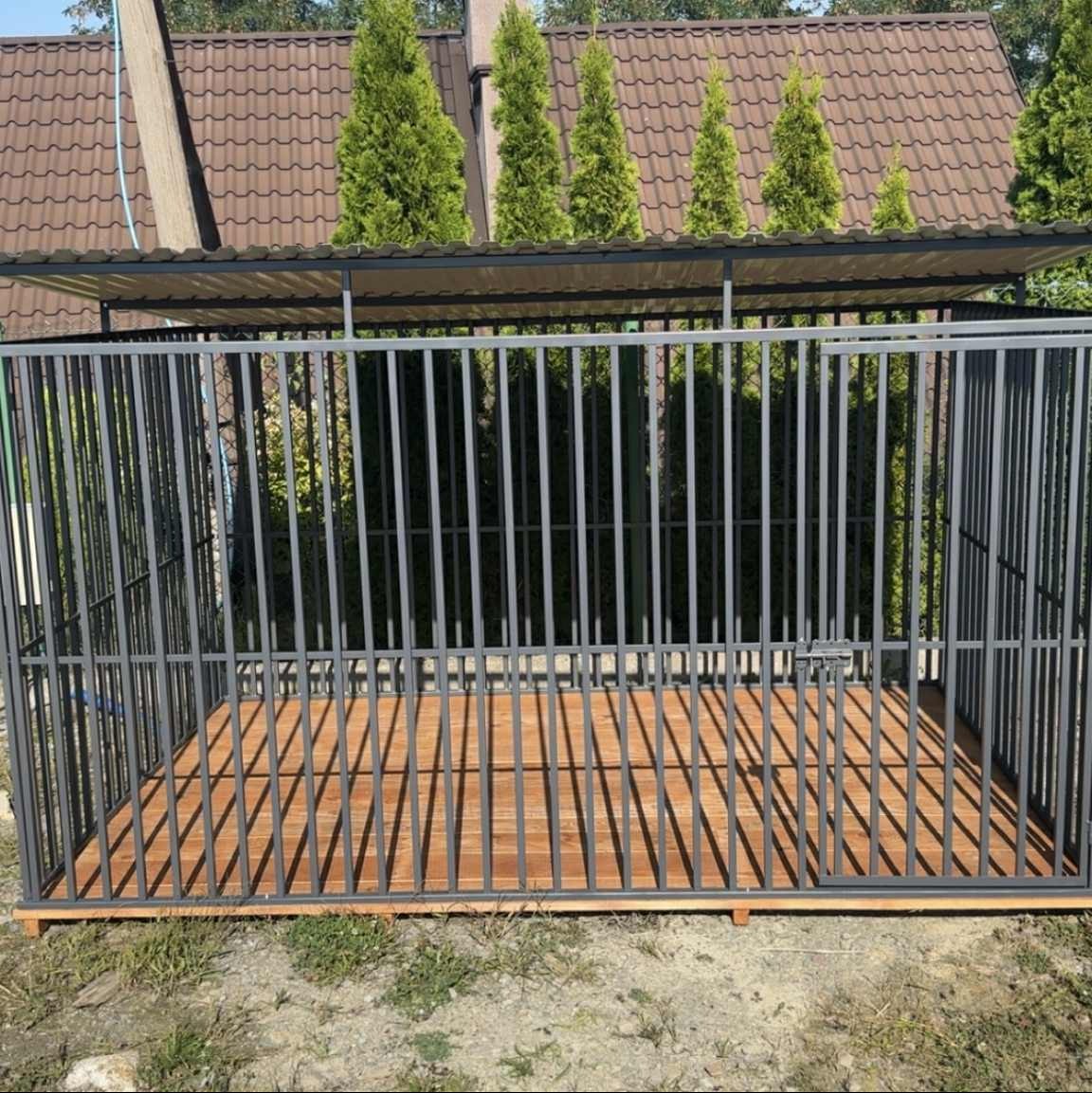 Metalowa konstrukcja zadaszonego kojca dla psa z drewnianą podłogą na zewnątrz, na tle zieleni i brązowego dachu domu.