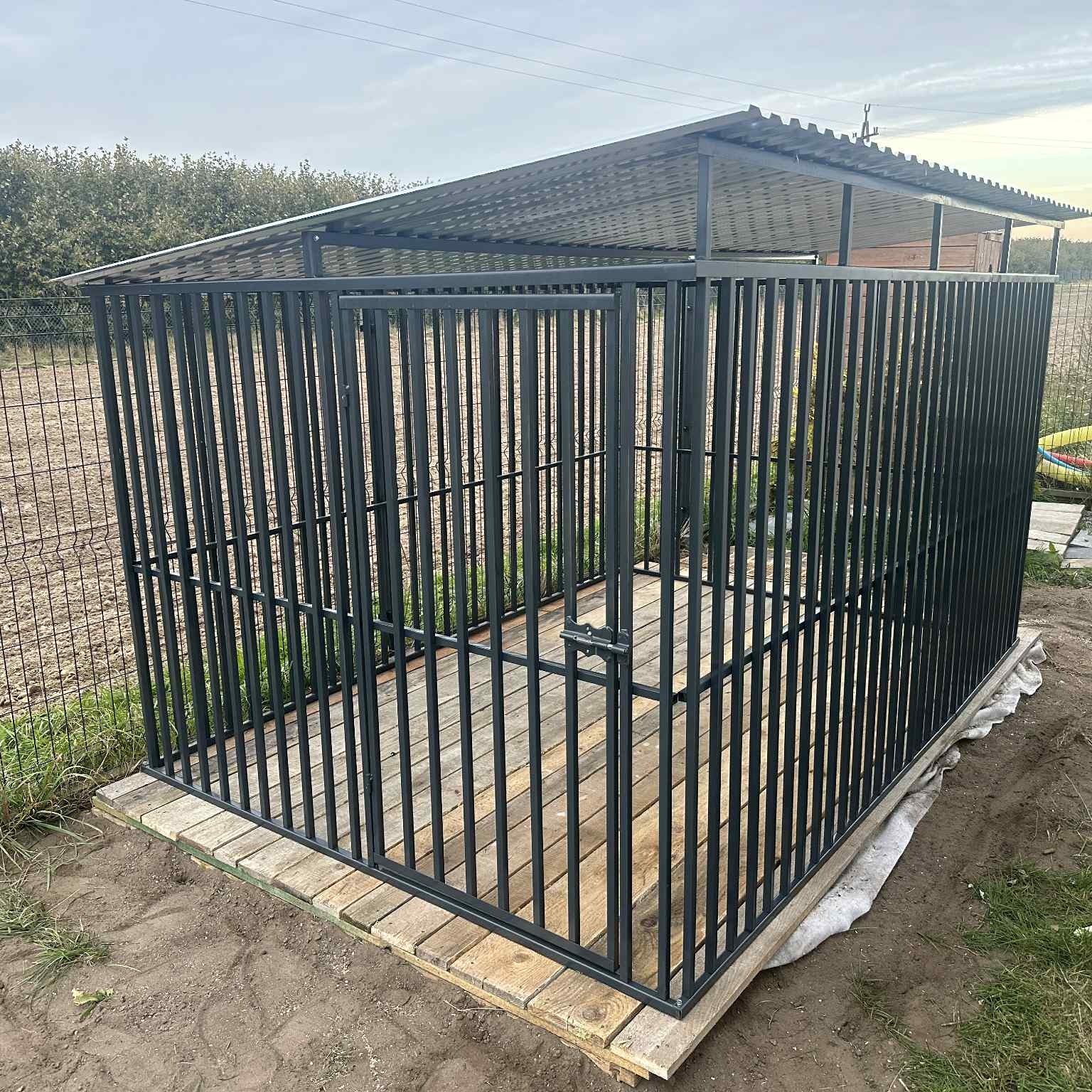 Metalowa konstrukcja z zadaszeniem, przypominająca klatkę lub kojec, na drewnianej podstawie, otoczona zielenią. Widoczne elementy zgrzewane i metalowe pręty.