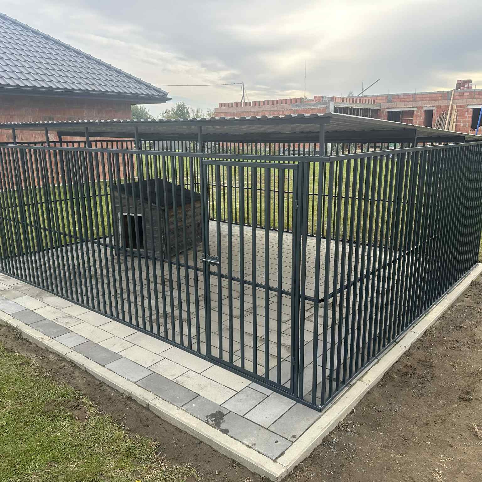 Metalowa konstrukcja z zadaszeniem i wybrukowanym podłożem, otaczająca budę dla psa. Widoczne elementy posesji w tle.