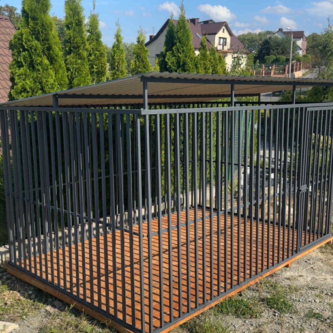 Metalowa konstrukcja z zadaszeniem i drewnianą podłogą, otoczona zielenią, na tle zabudowań mieszkalnych w słoneczny dzień. Widoczna brama wejściowa.