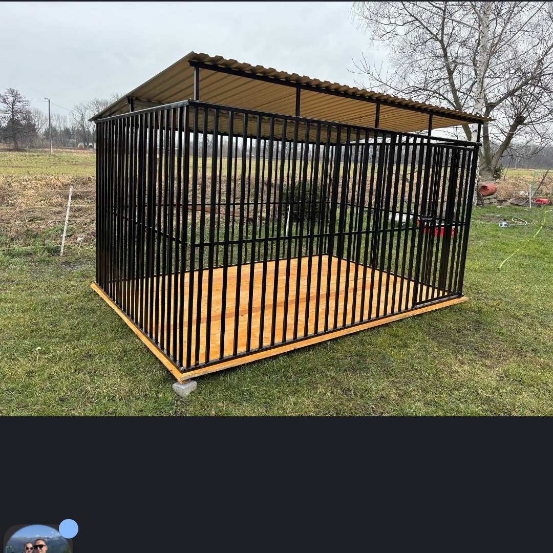 Metalowa konstrukcja z zadaszeniem na trawie, czarne pręty, drewniana podłoga, betonowe podstawy, widok z boku, pochmurne niebo w tle.