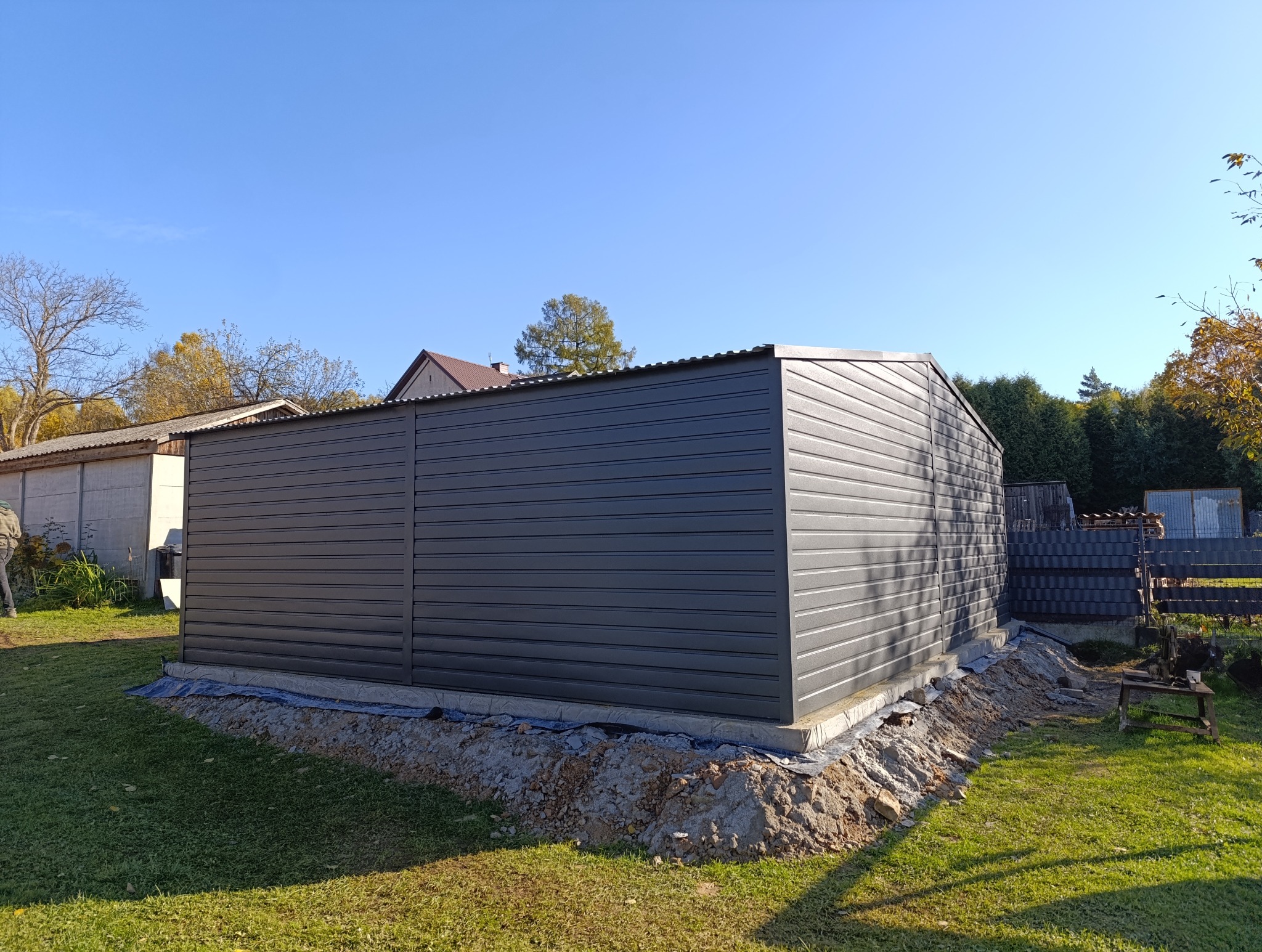 Szary garaż blaszany z poziomym wzorem paneli, widoczny na tle zielonego trawnika i błękitnego nieba. Ukończona konstrukcja na fundamencie, w tle budynki mieszkalne.