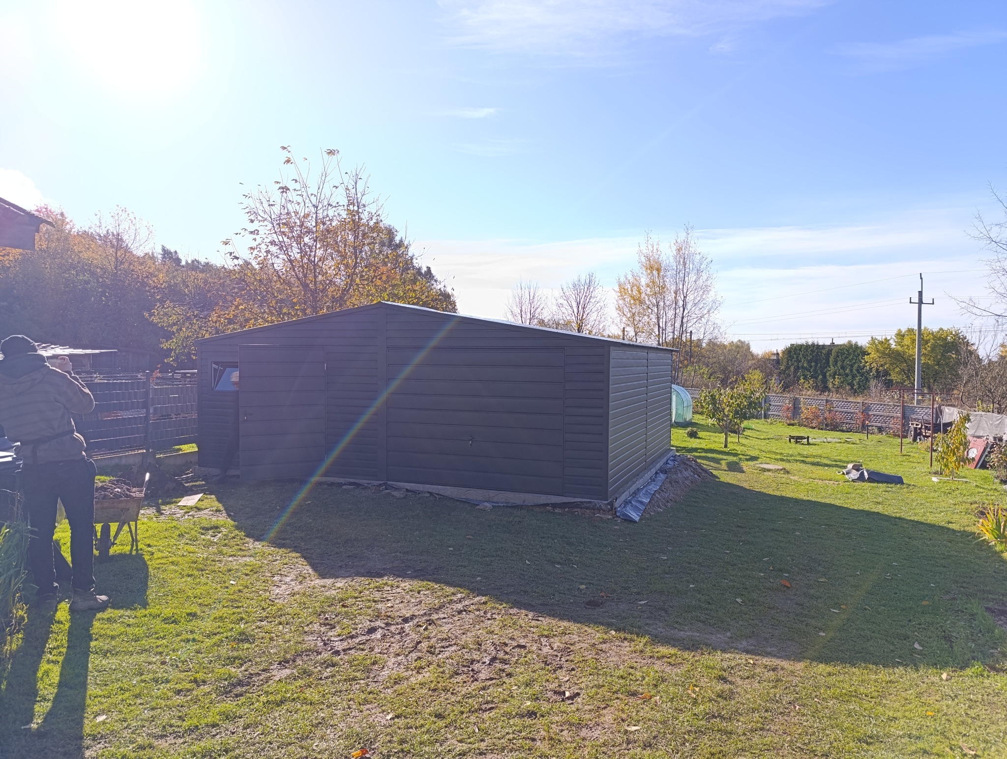 Nowoczesny, grafitowy garaż blaszany z bramą uchylną, usytuowany na trawniku w ogrodzie. Widoczny fragment osoby z taczką. Słoneczny dzień, błękitne niebo.