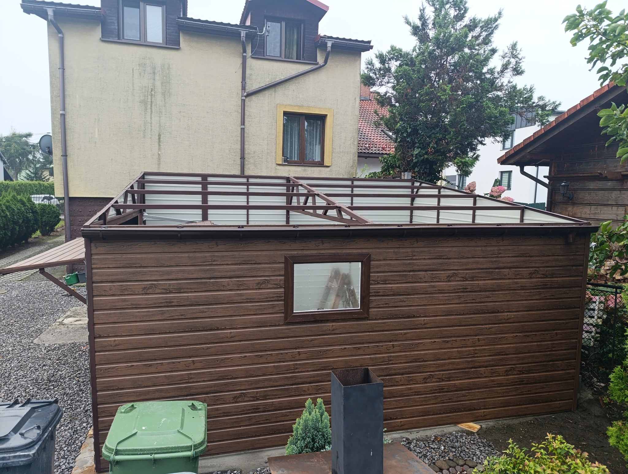 Brązowy garaż blaszany z oknem i częściowo zadaszoną konstrukcją dachu, widoczny na tle domu i zieleni. Ujęcie z poziomu gruntu, z widocznymi koszami na śmieci.