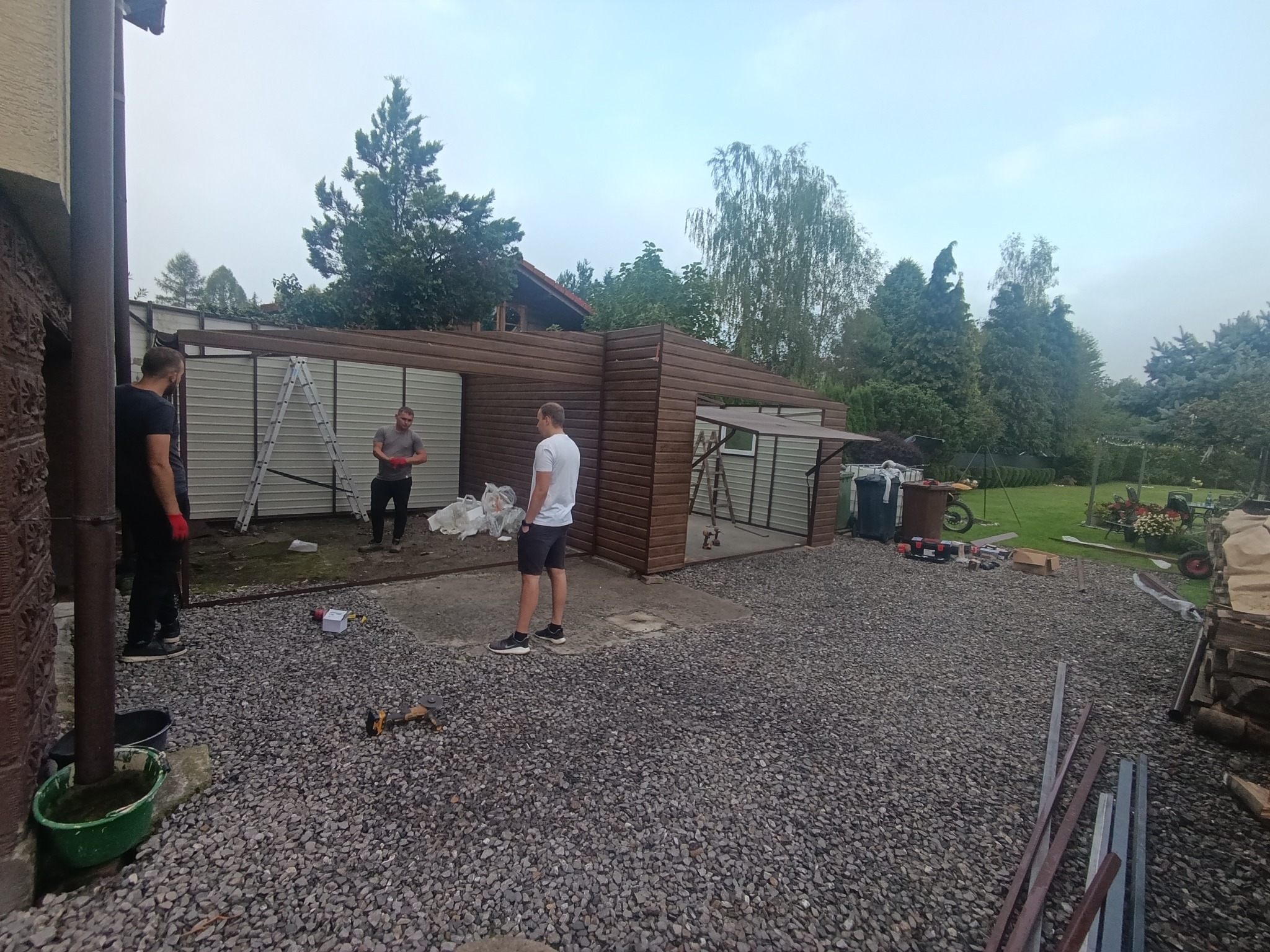 Montaż garażu blaszanego z brązowymi elementami wykończeniowymi. Widoczni trzej mężczyźni podczas pracy, drabina i narzędzia na żwirowym podłożu.