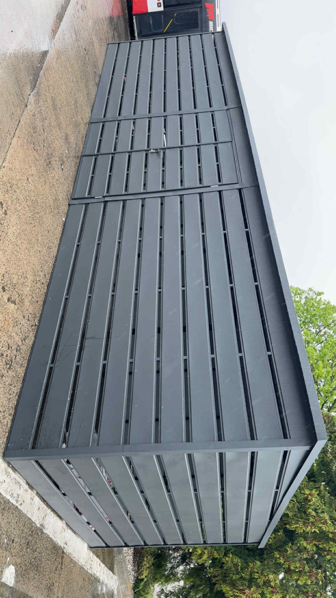 Nowoczesna, szara pergola metalowa z pionowymi lamelami, widziana pod kątem. W tle betonowa nawierzchnia i zieleń drzew. Minimalistyczny design.