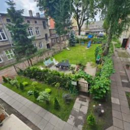 Widok z góry na zadbany ogród z placem zabaw, meblami ogrodowymi i bujną roślinnością, otoczony starymi budynkami mieszkalnymi. W tle widoczny samochód. Ogród oddzielony od chodnika niskim żywopłotem.