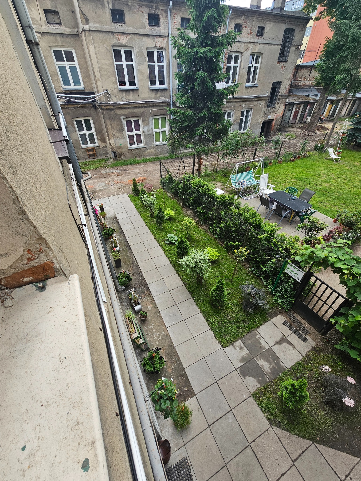 Widok z góry na zadbany ogród z roślinami ozdobnymi, ścieżką z płyt i meblami ogrodowymi, na tle starej kamienicy.