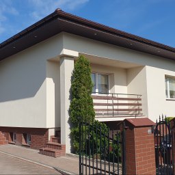 for your comfort - Jasna elewacja domu jednorodzinnego z balkonem, brązowym dachem i elementami z cegły klinkierowej, widok z poziomu ulicy przez czarne, kute ogrodzenie.