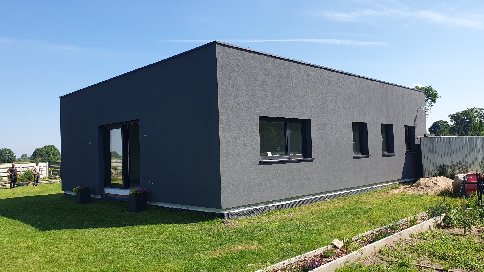 Nowoczesny, parterowy dom z grafitową elewacją o fakturze baranka, z trzema oknami i przesuwnymi drzwiami balkonowymi, otoczony trawnikiem i rabatami kwiatowymi w słoneczny dzień.