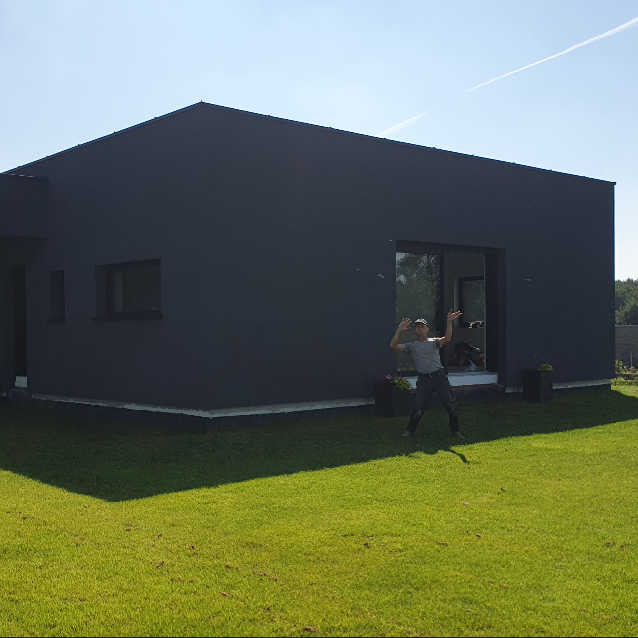 Nowoczesny, czarny dom jednorodzinny z płaskim dachem, zadowolony pracownik budowlany pozujący na tle budynku na tle zielonego trawnika.