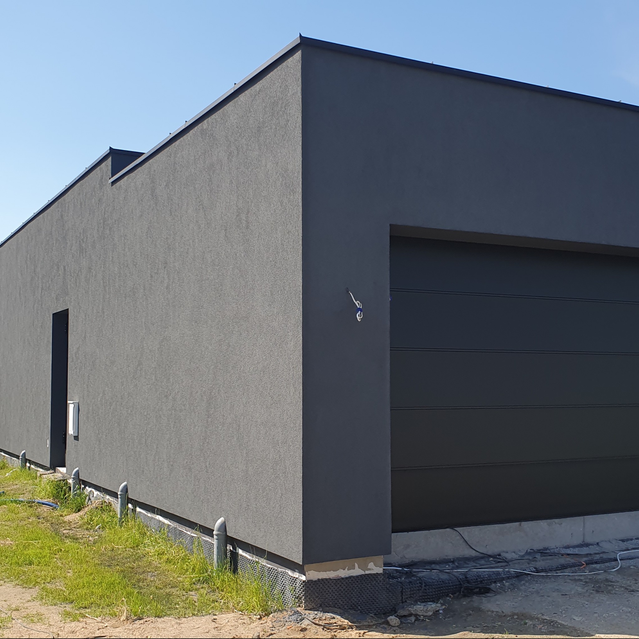 Nowoczesny, szary dom jednorodzinny z garażem i elewacją o wyraźnej fakturze, widoczne rury wentylacyjne przy fundamencie.