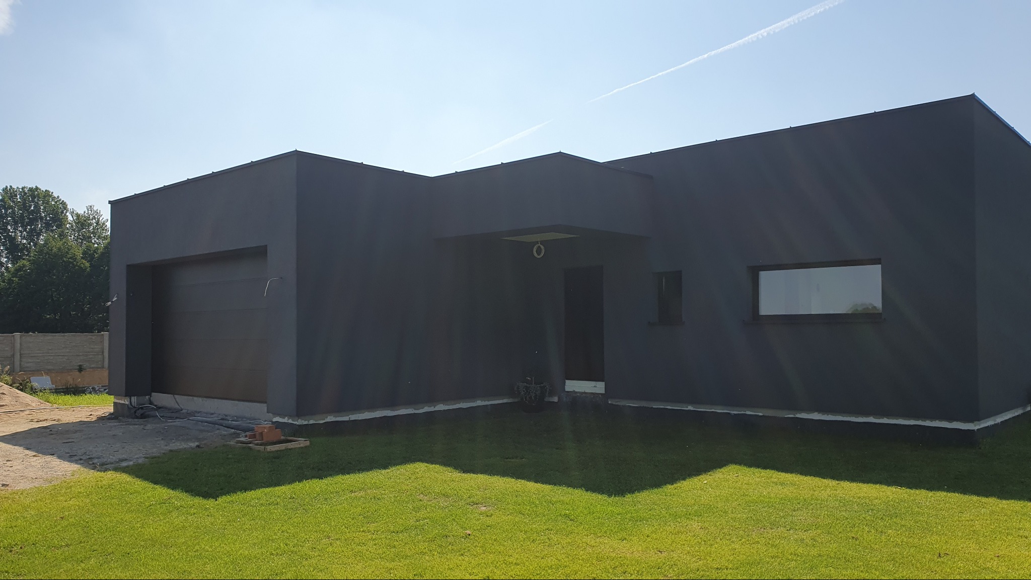 Nowoczesny, grafitowy dom jednorodzinny z płaskim dachem, widok z zewnątrz na elewację i garaż, trawnik przed budynkiem, błękitne niebo z smugą kondensacyjną samolotu.