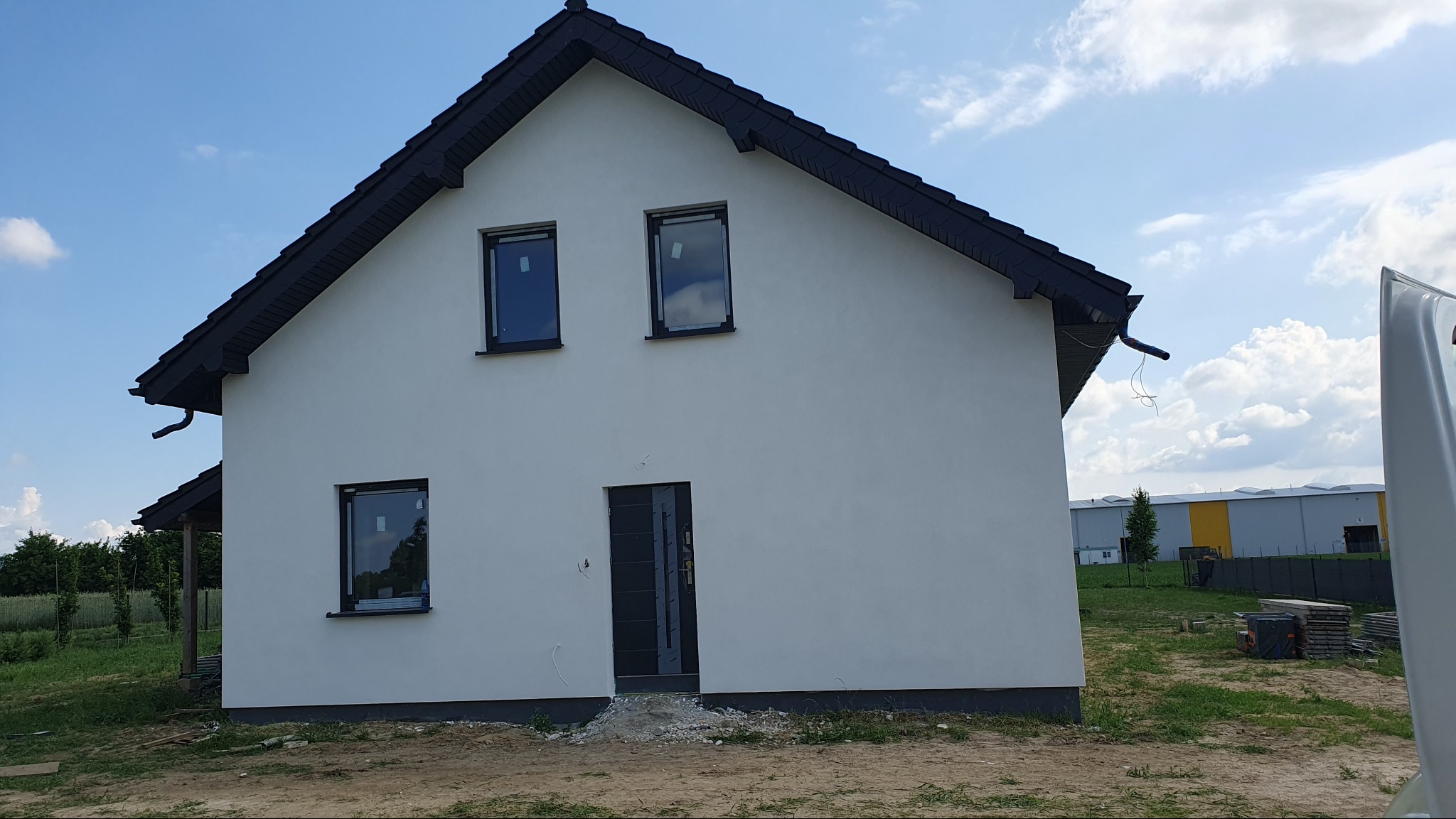 Nowo otynkowany dom jednorodzinny z czarnym dachem i stolarką okienną, widoczne niedokończone prace wykończeniowe wokół wejścia.