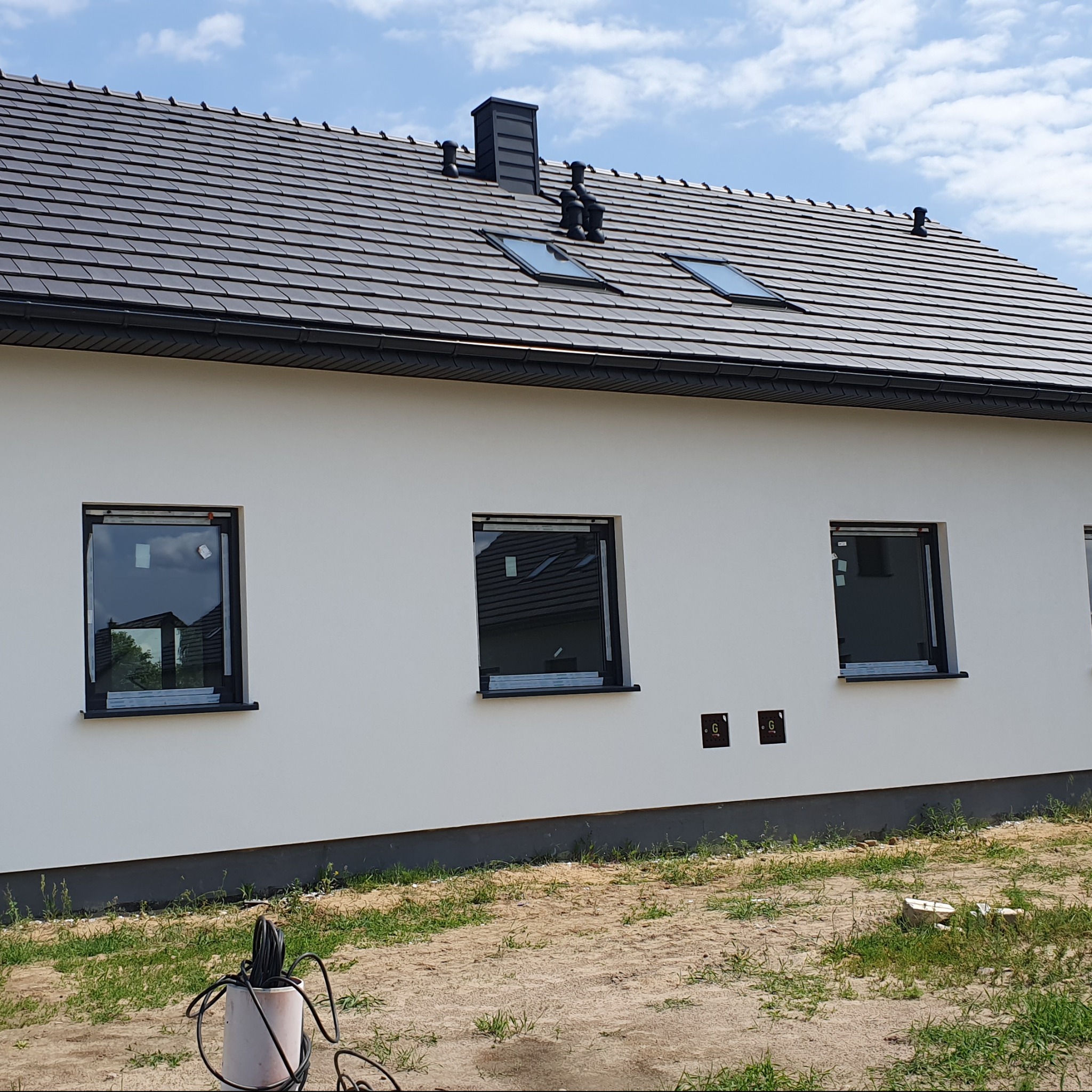 Nowoczesny dom jednorodzinny z białą elewacją, grafitowym dachem z dachówki ceramicznej, oknami w czarnych ramach, widoczne przyłącza na elewacji i teren wokół domu w trakcie zagospodarowywania.