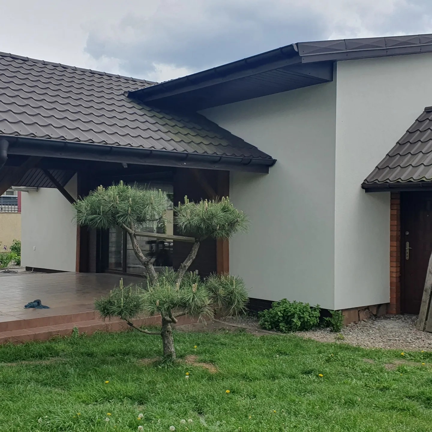 Jasna elewacja domu jednorodzinnego z brązowym dachem, widok z ogrodu z ozdobnym drzewkiem bonsai na pierwszym planie.