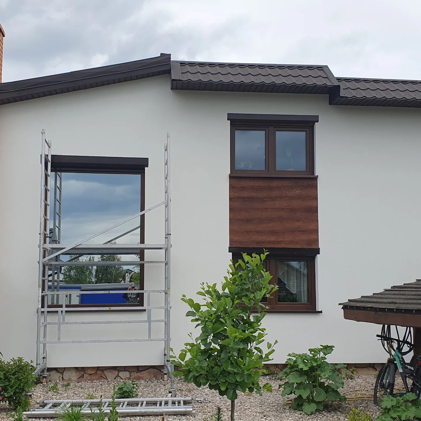 Biała elewacja domu jednorodzinnego z brązowymi oknami i fragmentem drewnianej okładziny, częściowo zasłonięta przez metalowe rusztowanie.