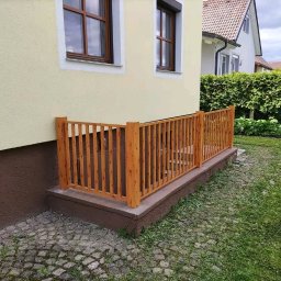 IronTech - Drewniane ogrodzenie zabezpieczające zejście do piwnicy w domu jednorodzinnym, widoczne z zewnątrz, z trawnikiem i kostką brukową wokół.