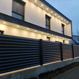 IronTech - Nowoczesne, poziome ogrodzenie z ciemnych paneli, oświetlony budynek w tle, widok z poziomu ulicy.