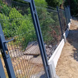 Nowoczesne ogrodzenie panelowe z metalową bramką, osadzone na betonowej podmurówce, widoczne z perspektywy bliskiej ziemi, z zielenią w tle.